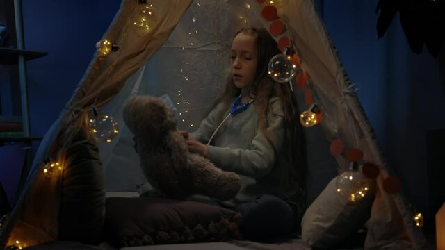 Teen girl wearing medical mask to teddy bear while sitting on floor in decorative makeshift tent at home. Child with toy stethoscope playing in evening. Concept of leisure.