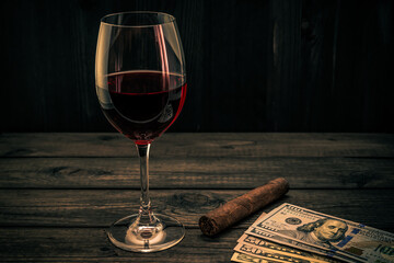 Glass of red wine with money and cuban cigar on an old wooden table. Focus on the cuban cigar