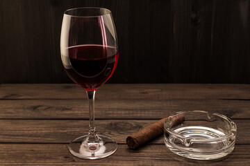 Glass of red wine and cuban cigar with glass ashtray on an old wooden table. Focus on the cuban cigar