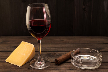 Glass of red wine with a piece of parmesan and cuban cigar with glass ashtray on an old wooden table. Focus on the cuban cigar