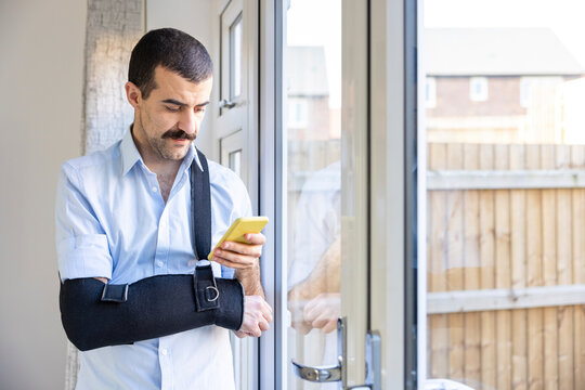 Man wearing arm sling using smart phone while standing by window at home