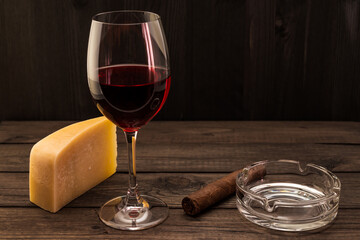 Glass of red wine with a piece of parmesan and cuban cigar with glass ashtray on an old wooden table. Focus on the cuban cigar