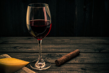 Glass of red wine with a piece of parmesan and cuban cigar on an old wooden table. Focus on the cuban cigar