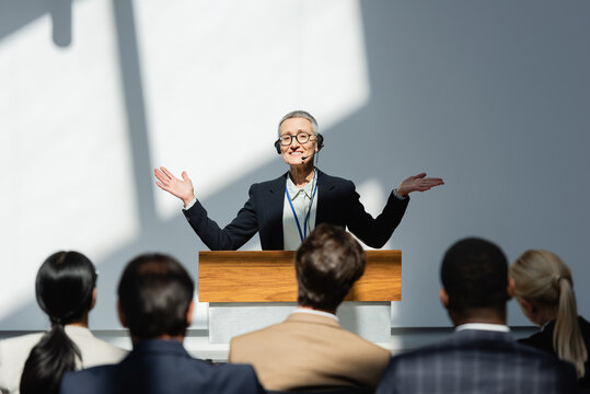 smiling lecturer standing with open arms during conference near participants on blurred foreground