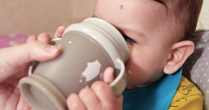 Cute baby boy drinking a glass of water at home. Close-up. The child drinks water from a plastic sippy cup