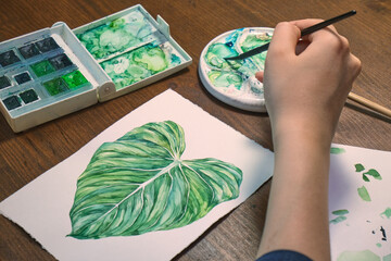 girl draws philodendron on paper in watercolor. mixes paints in a palette