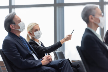 young businesswoman in medical mask pointing with pen near colleagues during seminar