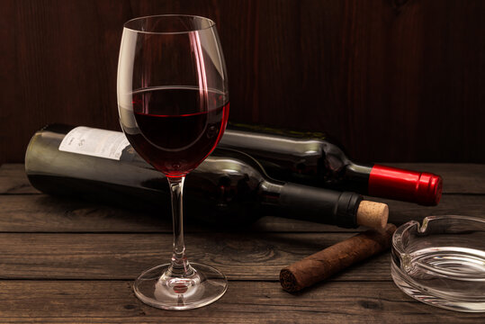 Two bottles of red wine with a glass and cuban cigar with ashtray on an old wooden table. Focus on the cuban cigar