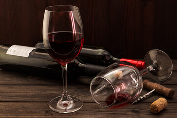 Two bottles of red wine with two glasses and corkscrew with cork on an old wooden table. Focus on an overturned glass of wine