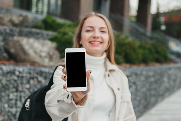 Happy smiling student girl showing mobile phone against the background of the college. Mockup for design