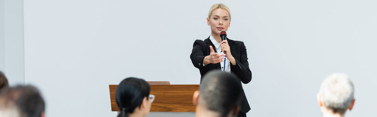 blonde speaker holding microphone while pointing at participants during seminar, banner