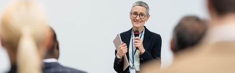 Mature businesswoman with microphone and paper folder talking near business people on blurred...