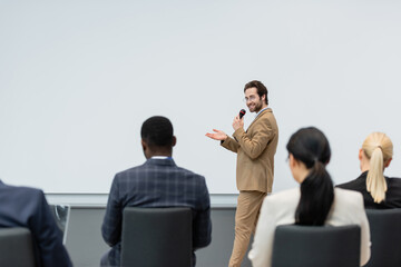 Smiling speaker talking and pointing with hand near blurred multiethnic business people during...