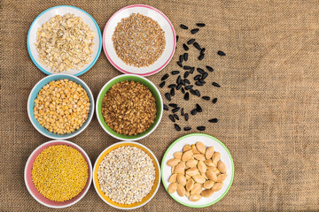 Portions of various cereals - wheat, peas, buckwheat, millet, oatmeal, barley in a colored dish against a background of burlap and scattered sunflower seeds and flakes