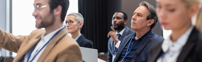 Middle aged businessman with laptop sitting near multiethnic colleagues during seminar, banner