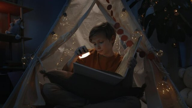 Young smart boy with blond hair in decorative tent reading interesting book at home in evening. Teenager sitting on floor while spending free time. Concept of leisure.