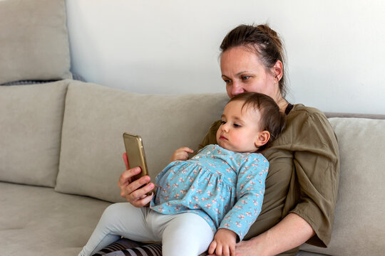 Single mother with baby girl having video call with family at home during the day. Caucasian mom and child playing games on the smartphone. Mother and baby playing with a smartphone sitting on a couch
