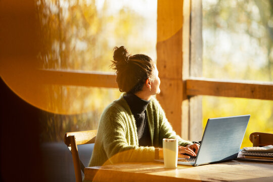 Woman working at laptop looking out window in sunny cafe