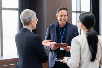 Smiling businessman with paper folder standing near colleagues with smartphone on blurred foreground