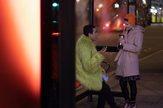 Stylish couple with coffee talking at bus stop in city at night