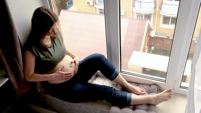 Beautiful smiling pregnant woman sitting near big window at home and caressing her belly. Pregnancy, motherhood and expectation of child concept.