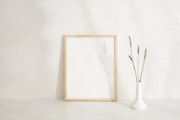 11x14 thin wood vertical frame mockup template resting against a neutral coloured wall. Summer sun shining through window creating artistic shadows. Artificial, dried flowers in a white vase.