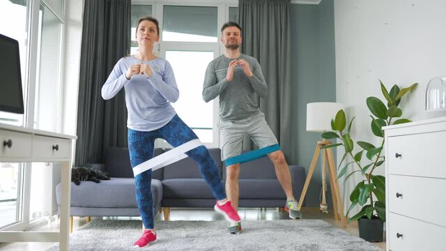 Caucasian couple is doing squats and leg swings with elastic band at home in cozy bright room, slow motion.