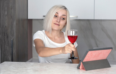 Beautiful young woman talking by video call via tablet, holding a glass of red wine