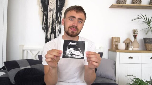 Pregnant man sitting on sofa at home and showing ultrasound image Baby scan