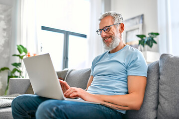 A mature man is sitting on the couch with a laptop on his lap at home. Freelance, work online. The man is texting on his laptop.