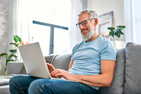 A mature man is sitting on the couch with a laptop on his lap at home. Freelance, work online. The man is texting on his laptop.