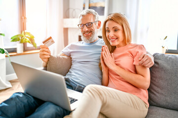 A mature couple is sitting on the couch at home with a laptop and a credit card in hand. Online shopping. Stay home concept.