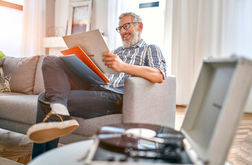 A mature man sits on the couch at home, relaxes, enjoys life and listens to vinyl records on a...