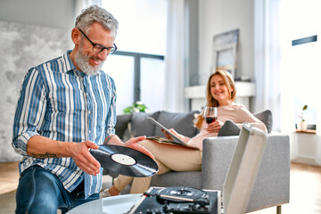 Mature couple are sitting at home on the couch, relaxing, enjoying life, drinking wine and listening to vinyl records on a music player. Romantic evening for a married couple.