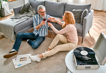 A mature couple are sitting on the floor near the sofa with glasses of wine, relaxing, enjoying life at home and listening to vinyl records on a music player. Romantic evening for a married couple.