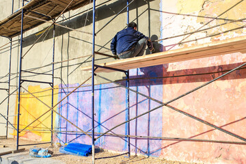 A worker paints the wall of a building while sitting on the scaffolding.