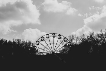 A fan-shaped Ferris wheel in the shade in the park and a light cloudy sky above it. Black and white atmospheric photo carousel in the park