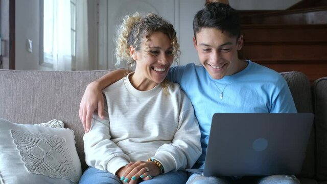Adult son and middle-aged 40s mother sitting on couch in cozy living room holding on lap notebook laughing on funny comedy movie, enjoy on-line amusements websites, internet virtual fun online concept