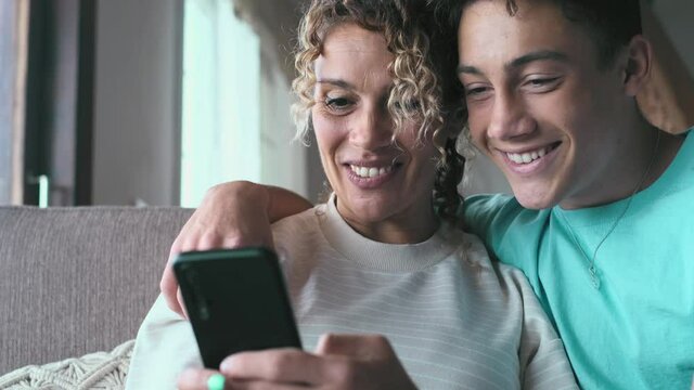 Smiling middle-aged 40s mother rest with grown-up son using smartphone together, happy young man enjoy family weekend with mom browsing wireless Internet on cellphone, have fun at home
