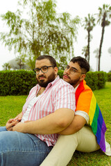 Retrato frontal de pareja de novios gay con barba abrazándose en el parque con una bandera LGBTQ 