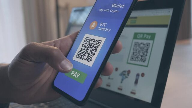 Close up of an African-American male hand holding a mobile phone, scanning QR code on a laptop monitor and paying with bitcoins payment mobile app