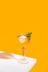Elegant stemware glass of fresh orange cocktail with big ice ball on white table surface, bright yellow background