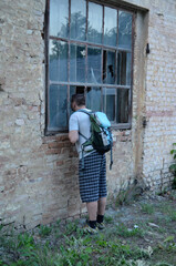 Obraz na płótnie Canvas A man with a backpack on his back looks through a broken window into an abandoned house