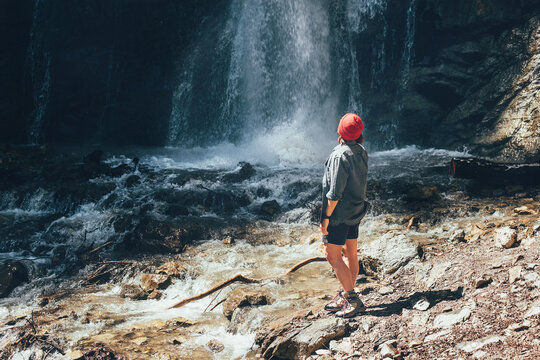 Woman dressed active trekking clothes standing near the mountain river waterfall and enjoying the splashing Nature power. Traveling, trekking and nature concept image.