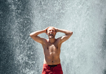 Middle-aged topless sincerely laughing man standing under the mountain river waterfall and enjoying the splashing Nature power. Fit people, trekking, and a natural beauty concept image.