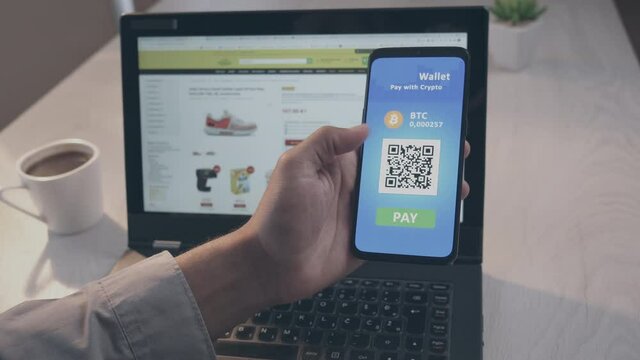 POV shot of a man sitting at the desk, holding a smartphone in the left hand, bringing it closer to a laptop screen, scanning QR code and paying with crypto wallet mobile app