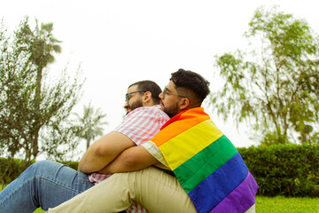Pareja de novios gay con barba abrazándose en el parque con una bandera LGBTQ 