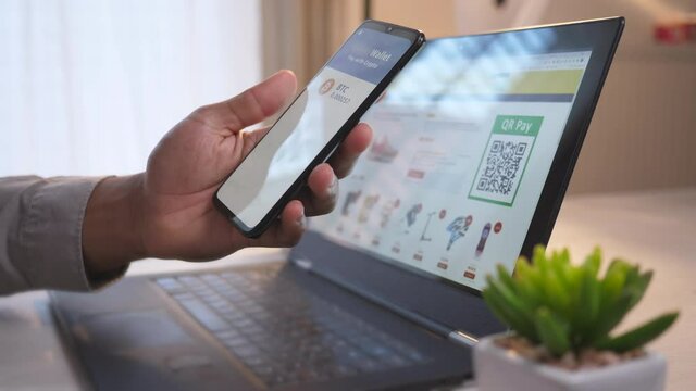 Handheld shot of an African-American male hand holding a mobile phone, using together with a laptop for online shopping, scanning QR code and paying with bitcoins via the mobile app