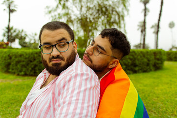 Pareja de novios gay con barba abrazándose y mirándose el uno al otro en el parque con una bandera LGBTQ  - obrazy, fototapety, plakaty