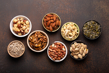 Selection of assorted raw nuts and various seeds in bowls on brown stone background from above, healthy source of energy, fat and vegetarian protein